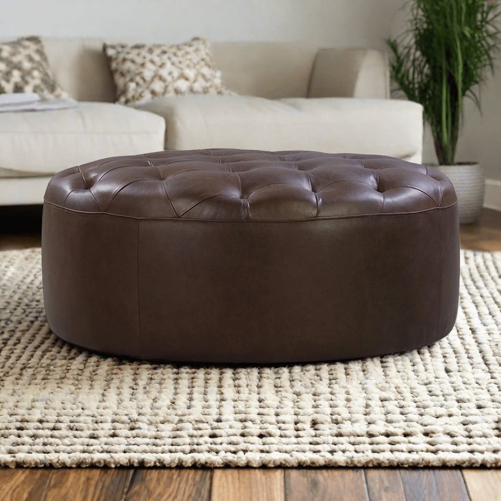 A round, tufted leather ottoman in a rich brown color, placed on a cream-colored, textured rug in a cozy living room setting with a beige couch and green plant in the background.