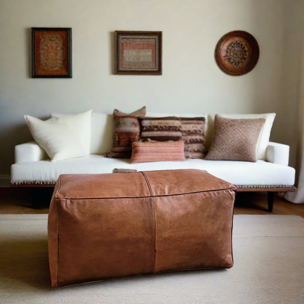 Handcrafted rectangular tan leather ottoman in a bohemian-style living room, highlighted by a white couch with various textured pillows and traditional wall art.