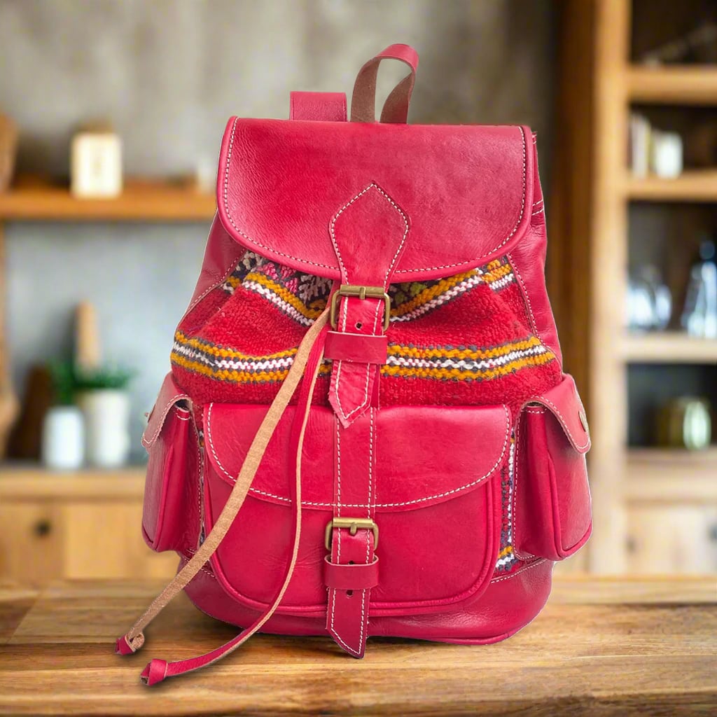 Front view of a handcrafted red leather backpack with vibrant kilim accents, featuring a buckle-closure flap, a large front pocket, and two snap-button side pockets, placed on a wooden table with rustic decor in the background