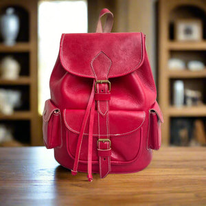 Front view of a handcrafted red Moroccan leather backpack featuring a top flap with a buckle closure, two snap-button side pockets, a large front pocket with a buckle strap, and smooth hand-stitched leather details, displayed on a wooden table with bookshelves in the background