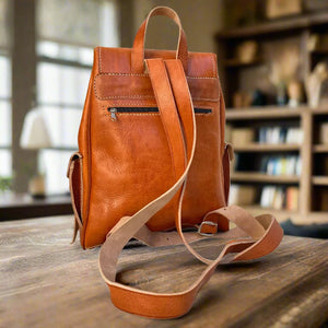 Back view of a handcrafted tan leather backpack featuring a zippered pocket and adjustable straps, placed on a wooden table with a cozy room in the background