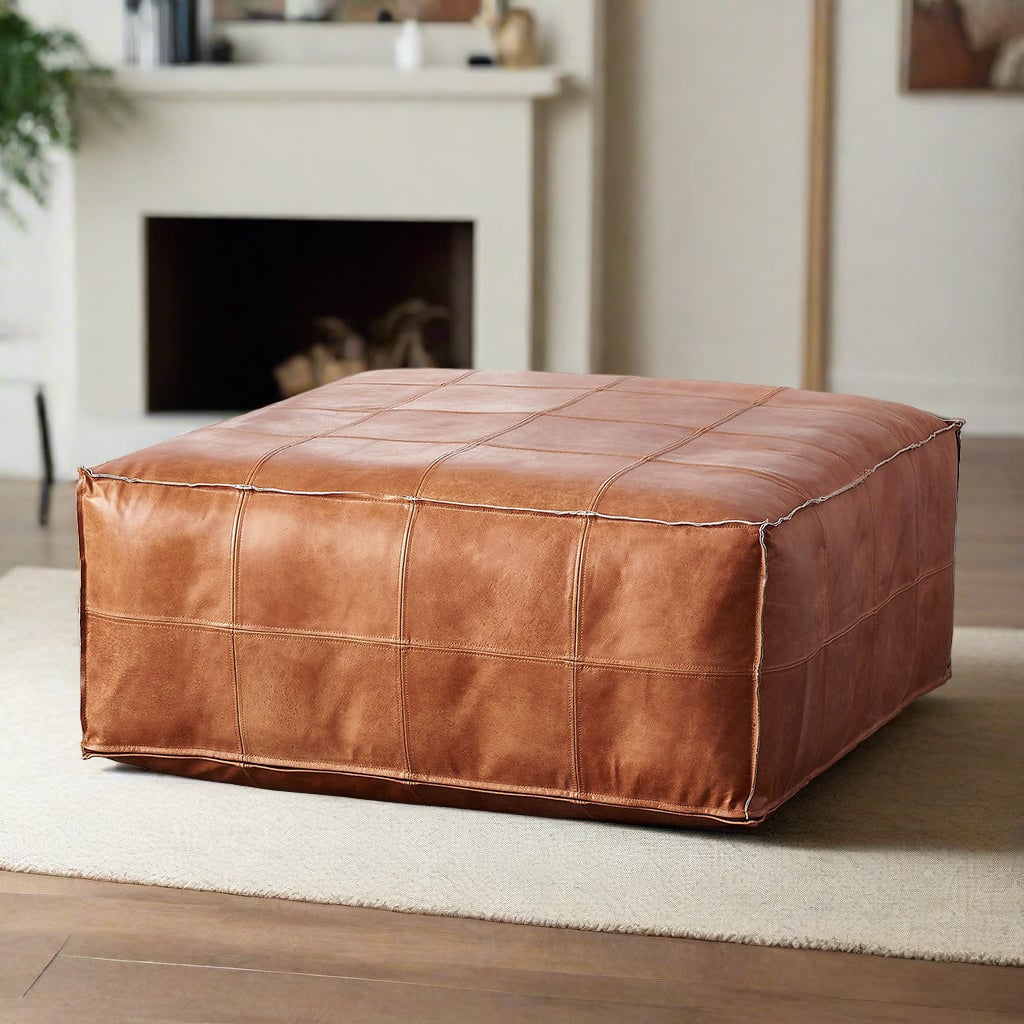 Handcrafted square tan leather ottoman in an elegant living room setting with a beige rug and a modern fireplace in the background.