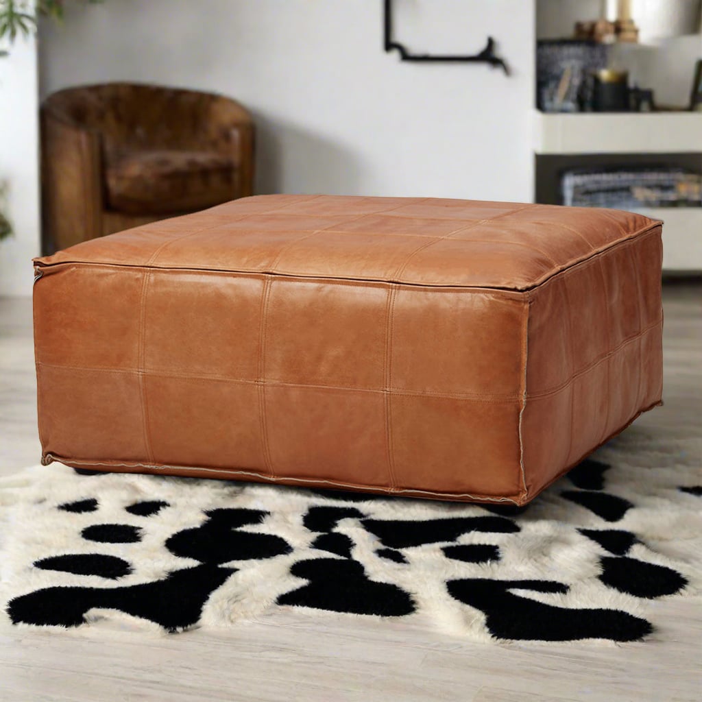 Handcrafted square tan leather ottoman on a black and white patterned rug in a stylish living room with contemporary decor.