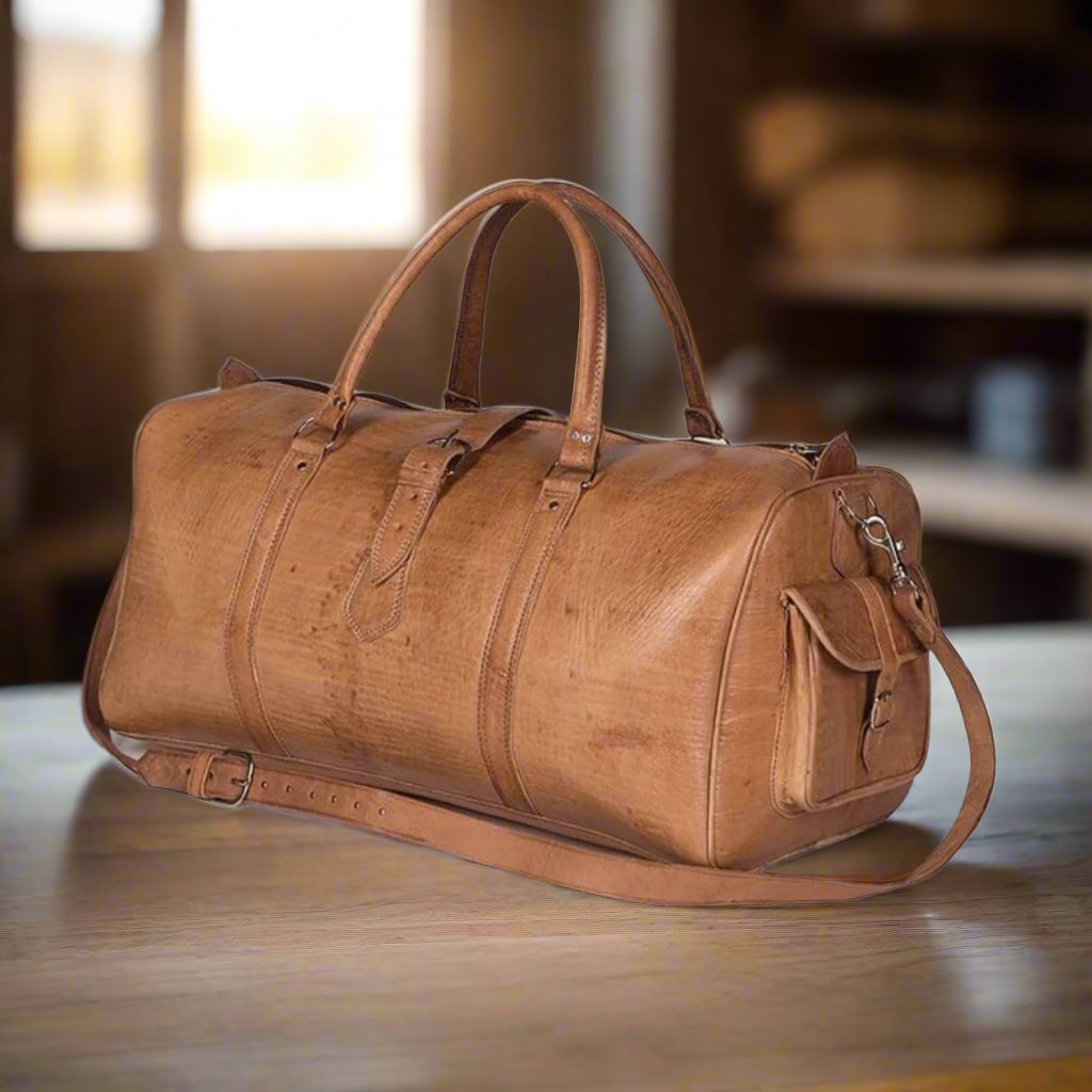 A handcrafted tan leather travel bag displayed on a wooden surface, featuring a smooth finish, reinforced handles, an adjustable shoulder strap, and a side pocket with a buckle closure. Perfect for weekend getaways or daily use.