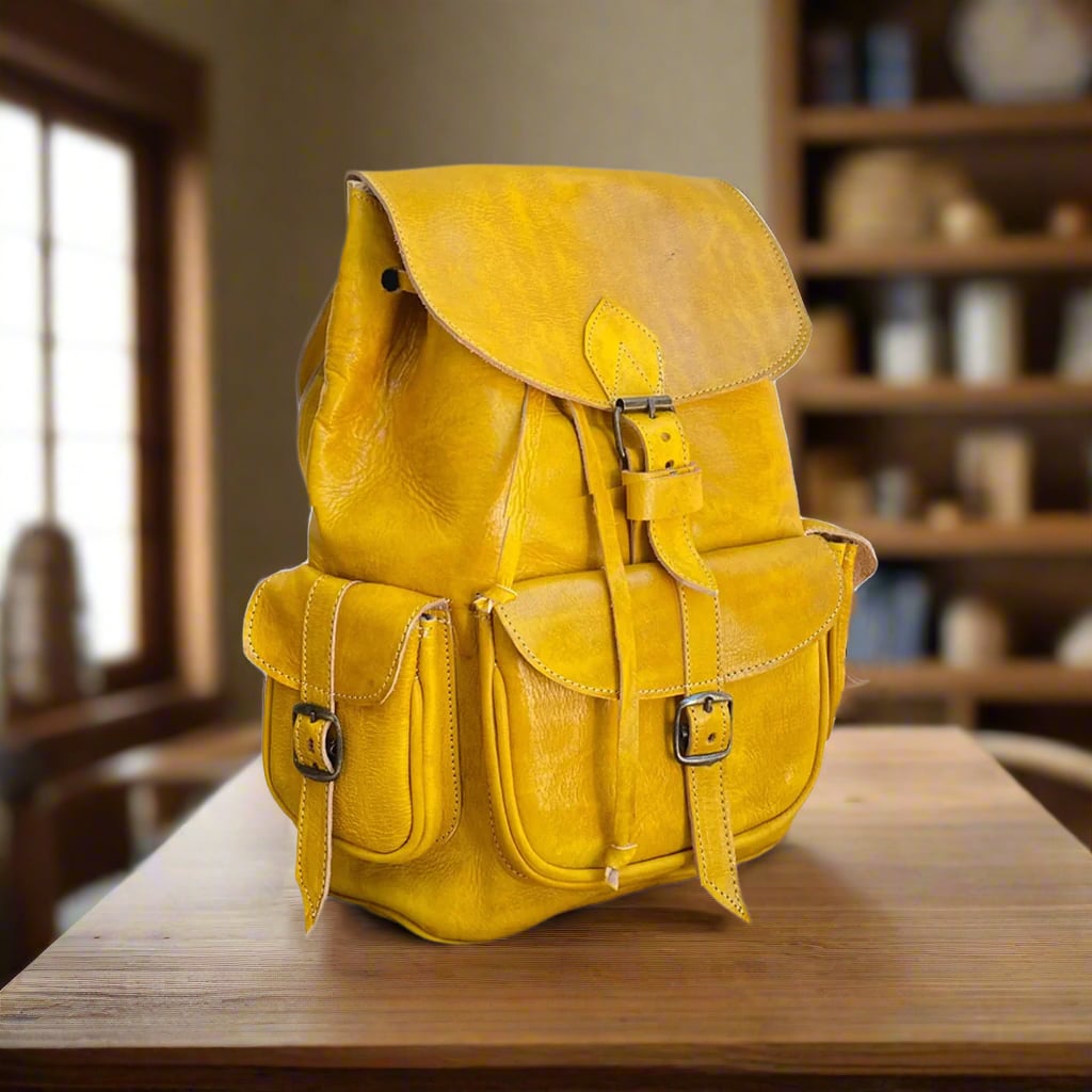 Handcrafted yellow Moroccan leather backpack with buckle closures and spacious front pockets, placed on a wooden table in a cozy room with bookshelves in the background