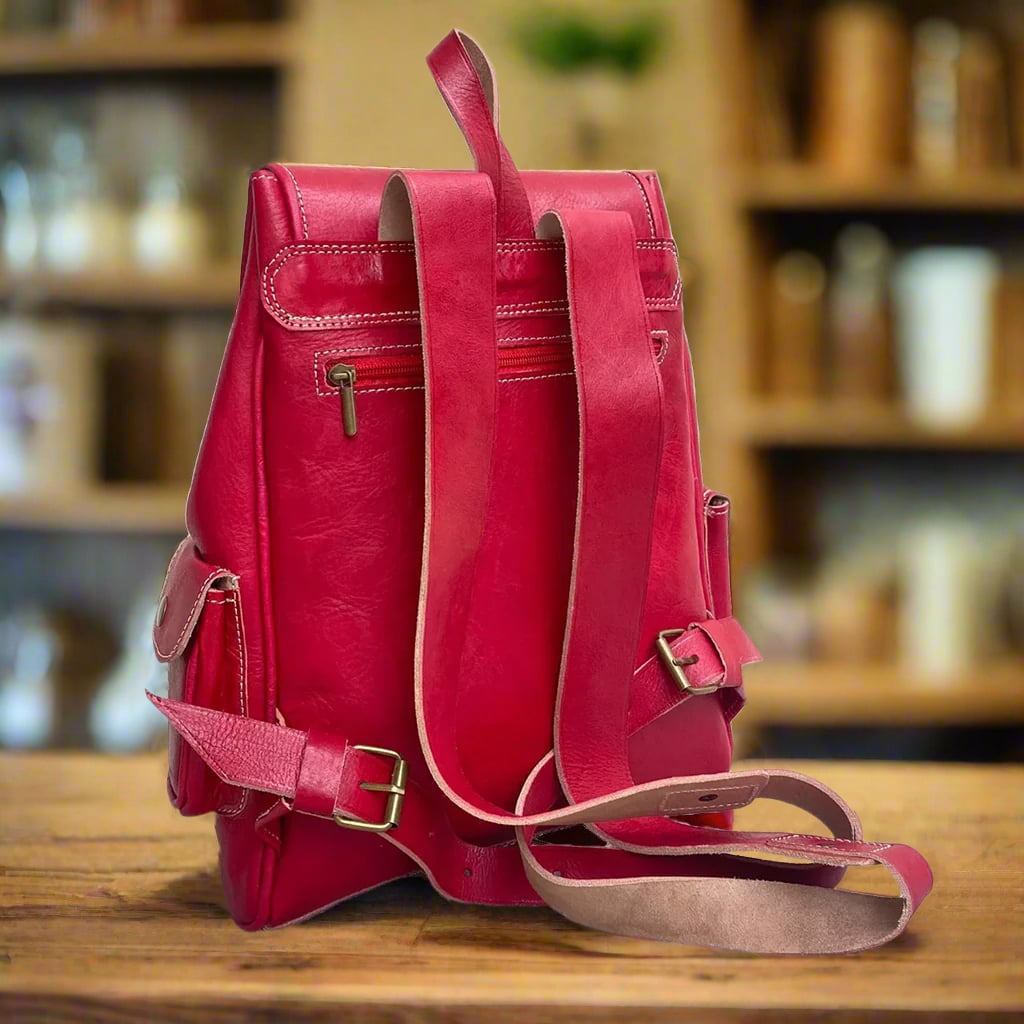 Back view of a red Moroccan leather backpack with adjustable leather straps, a secure zippered back pocket, detailed stitching along the edges, and reinforced seams, placed on a wooden surface in a cozy room with shelves and jars in the background