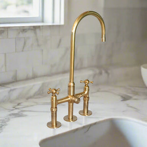 Tall gooseneck brass faucet with classic cross handles on a sleek marble kitchen worktop, positioned beside an under-mount sink and window