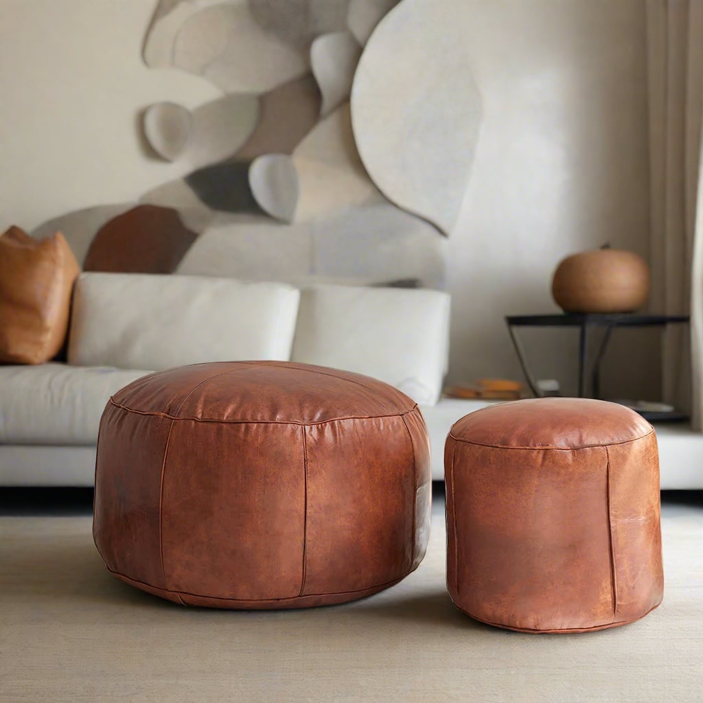 Two Moroccan leather poufs, one large and one small, both handcrafted with natural leather and detailed stitching, arranged together in a stylish living room with modern furnishings.