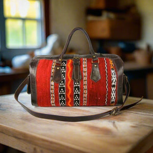 A handcrafted leather weekend bag featuring vibrant red, orange, and white handwoven kilim patterns, complemented by rich brown leather trim and sturdy handles. The bag sits on a rustic wooden table, with an adjustable leather shoulder strap neatly placed beside it.