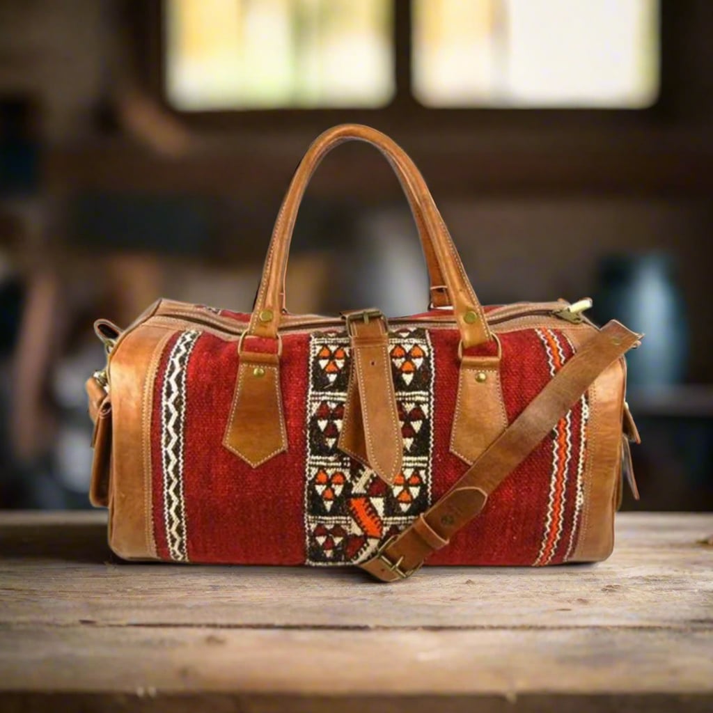 A handcrafted leather and kilim duffle bag featuring vibrant red panels with intricate geometric patterns, complemented by premium tan leather accents, sturdy handles, and an adjustable shoulder strap, resting on a wooden surface in a warm, rustic setting