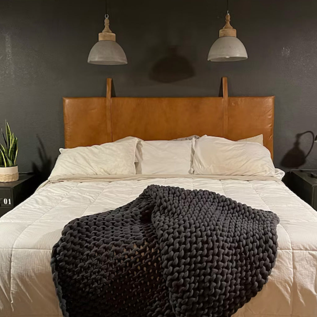 King-size bed with dark gray knit blanket, white sheets, pillows against dark wall with hanging leather headboard, selling lamps - Moroccan Interior