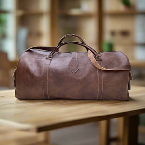 A minimalist brown leather duffle bag with durable stitched handles, an adjustable shoulder strap, and a spacious interior. The bag is displayed on a wooden table in a warm, cozy setting, showcasing its timeless and practical design