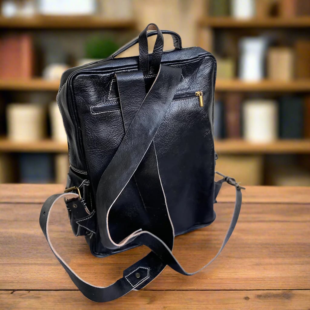 Back view of a black leather backpack with adjustable leather straps, a grab handle, and a zippered pocket, set against a warm-toned background.