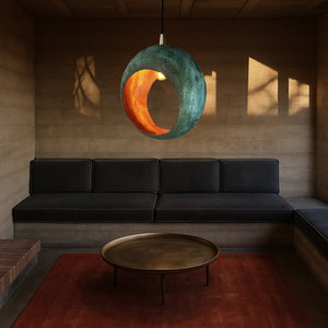 Modern minimalist living room with a dark sectional sofa, round brass coffee table, and a unique spherical pendant light featuring a green and orange patina finish, casting warm ambient lighting.
