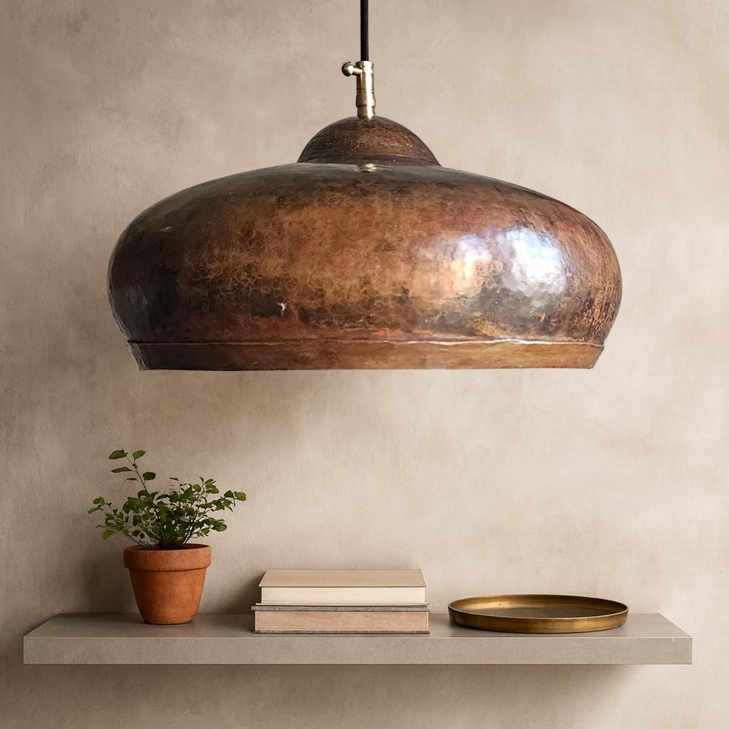 A handcrafted Moroccan pendant light in rustic hammered copper, hanging above a minimalist floating shelf with books, a plant, and a brass tray