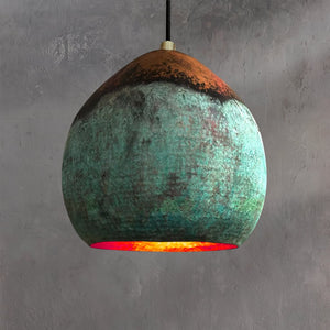 Handcrafted Moroccan pendant light with a rounded organic shape, featuring a vivid ombré patina blending copper and turquoise tones, glowing warmly against a textured gray background