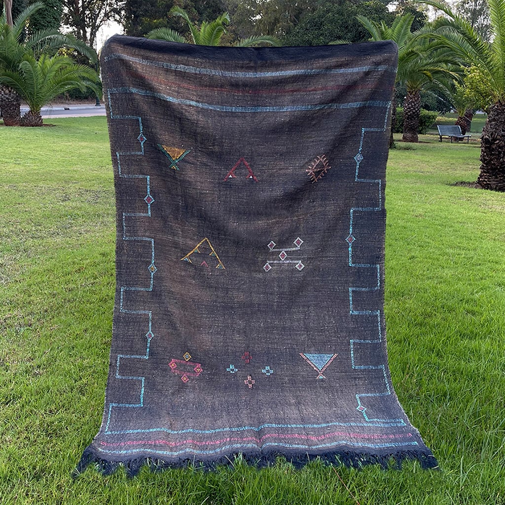 Handwoven black Moroccan sabra cactus silk rug with colorful Berber tribal motifs and fringed edges, displayed outdoors on green grass