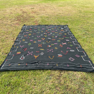 Handwoven Moroccan sabra cactus silk rug in black, decorated with colorful tribal embroidery patterns, laid flat on grass outdoors