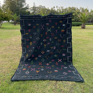Handwoven Moroccan sabra cactus silk rug in deep black with colorful geometric tribal embroidery, displayed outdoors on grass in a garden setting