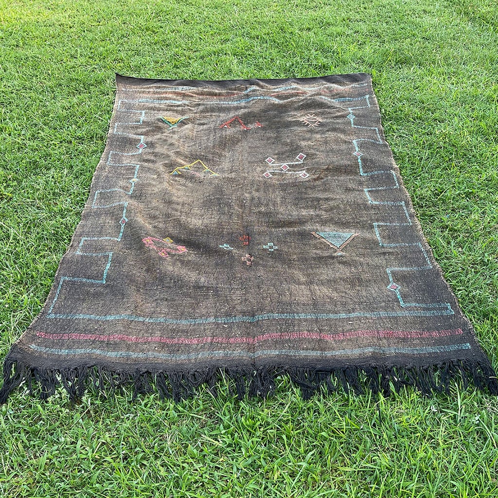 Black Moroccan sabra cactus silk rug laid flat on grass, featuring colorful embroidered Berber motifs, geometric tribal symbols, and fringed edges