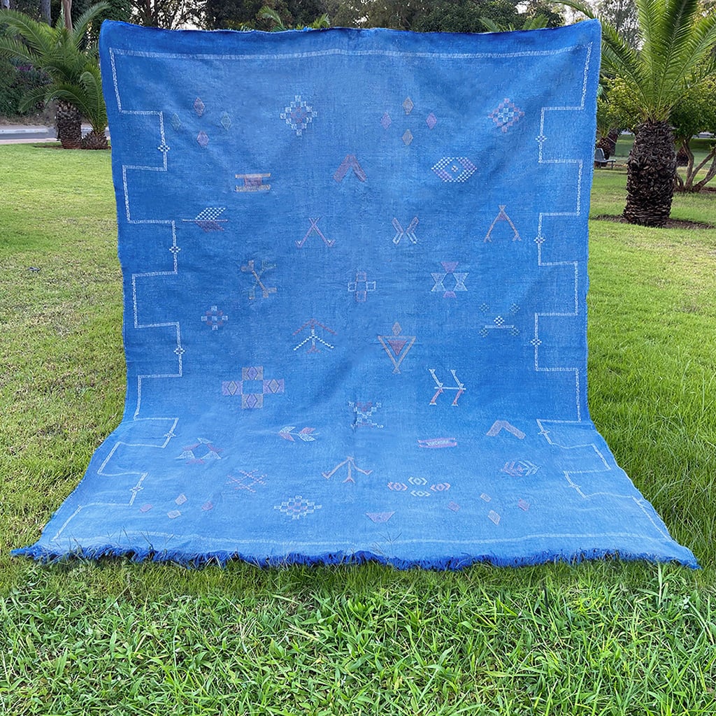 Flat-laid blue Moroccan sabra cactus silk rug on grass, featuring colorful Berber tribal motifs, geometric embroidery, and fringed edges