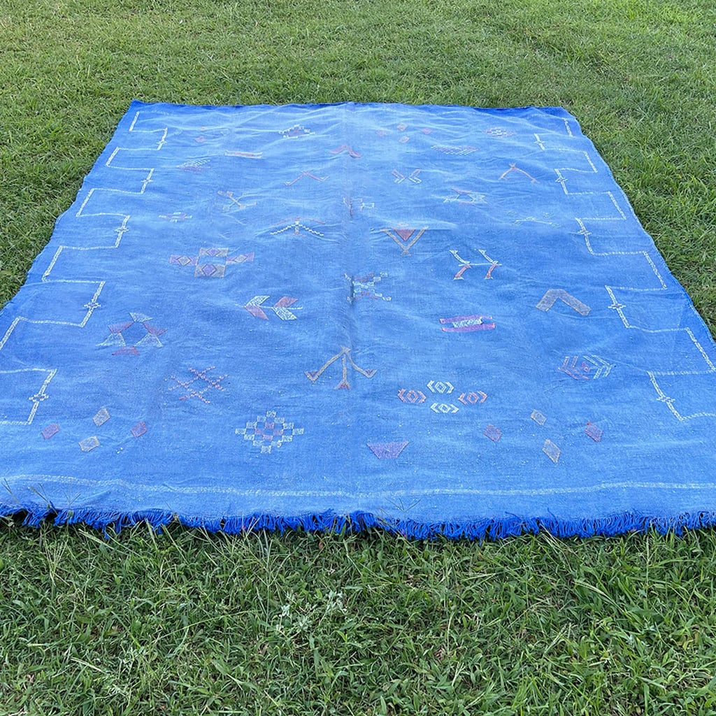 Blue Moroccan sabra cactus silk rug laid flat on grass, decorated with colorful Berber tribal motifs, geometric embroidery, and fringed edges