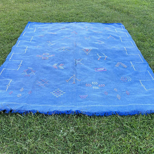 Blue Moroccan sabra cactus silk rug laid flat on grass, decorated with colorful Berber tribal motifs, geometric embroidery, and fringed edges
