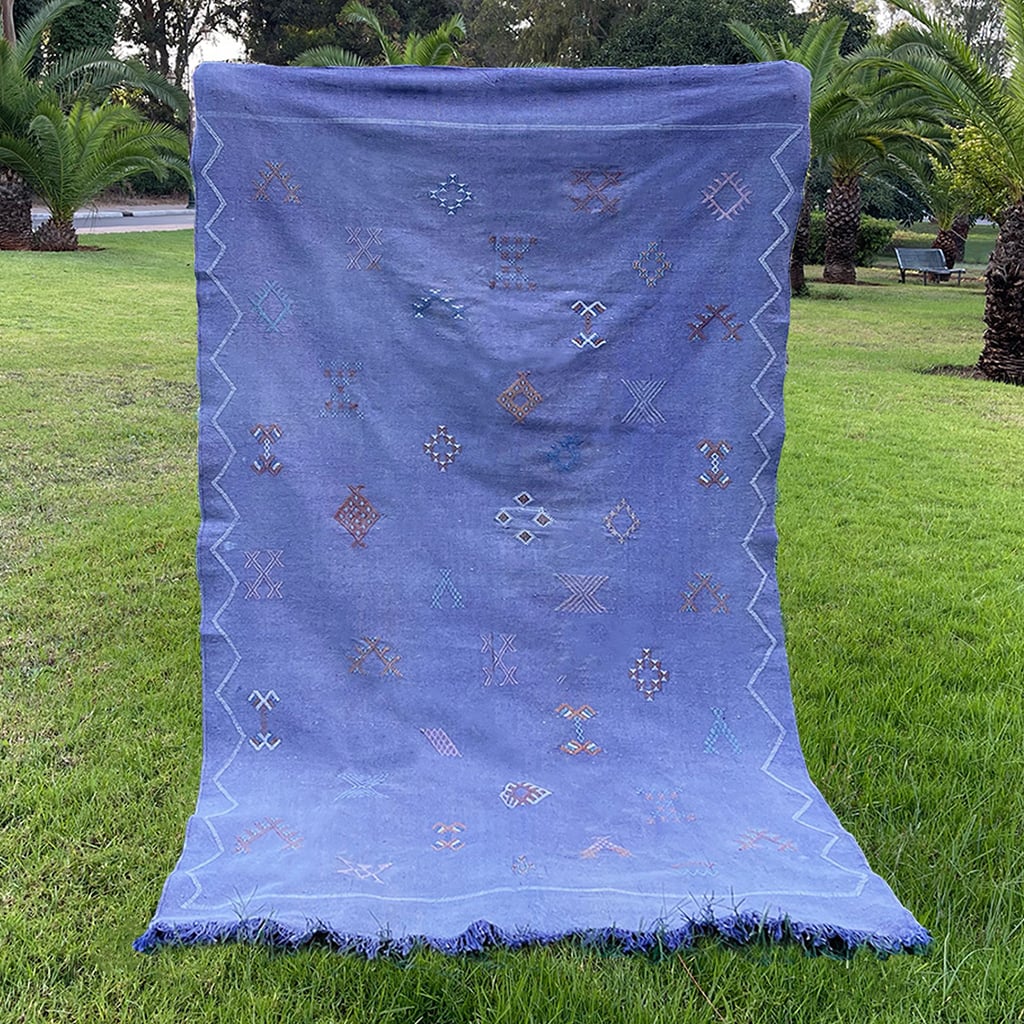 Handwoven Moroccan sabra cactus silk rug in indigo blue, featuring traditional tribal embroidery patterns and fringe edges, displayed outdoors on grass