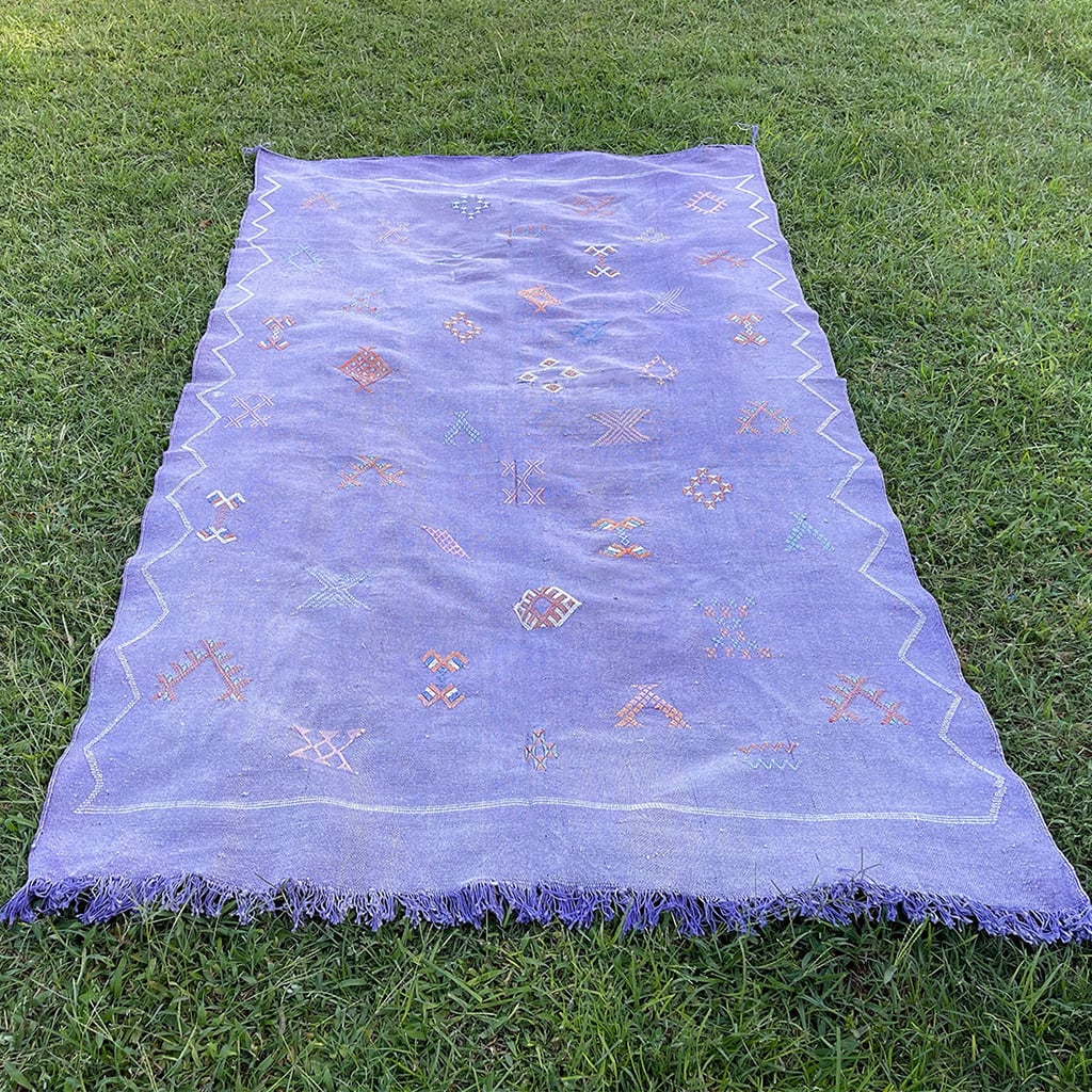 Handwoven Moroccan sabra cactus silk rug in soft lavender purple with traditional tribal embroidery and fringed edges, laid flat on green grass