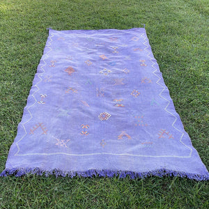 Handwoven Moroccan sabra cactus silk rug in soft lavender purple with traditional tribal embroidery and fringed edges, laid flat on green grass