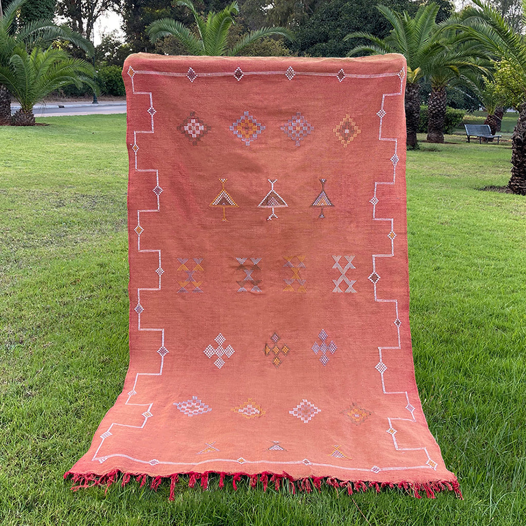 Handwoven Moroccan sabra cactus silk rug in terracotta with geometric tribal embroidery and red fringe, displayed on grass