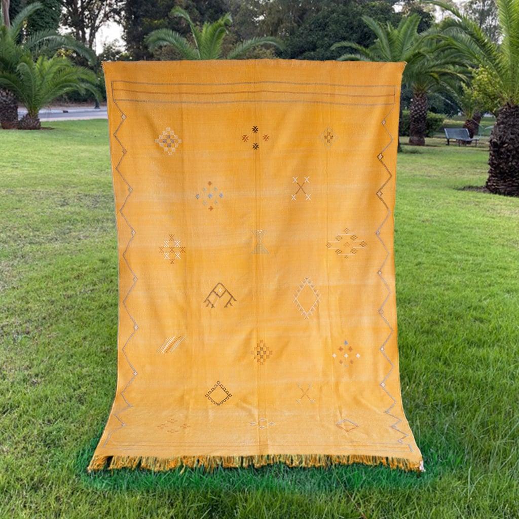 Bright mustard yellow Moroccan rug displayed on grass, featuring handwoven Berber symbols and geometric border details with orange fringe ends