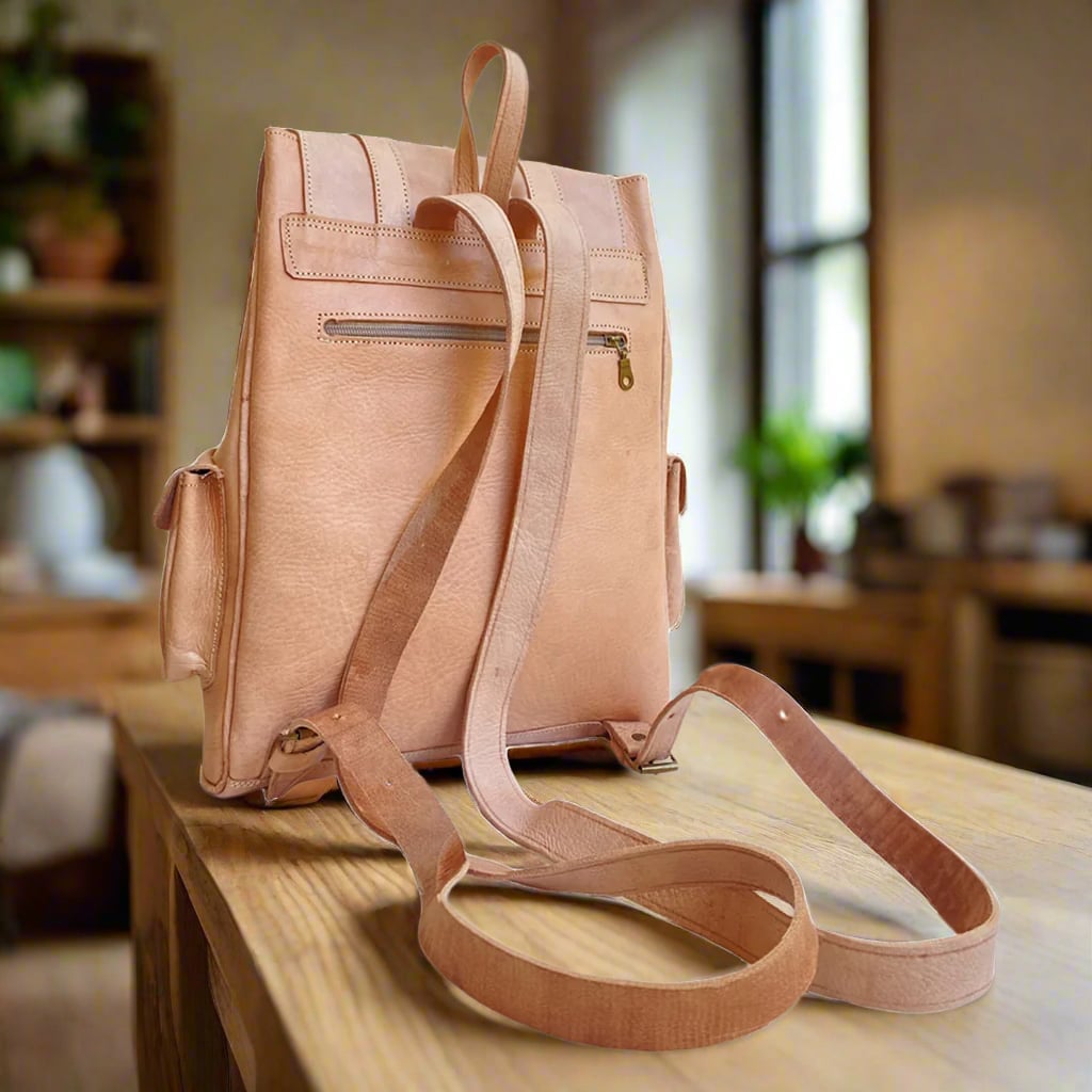 Back view of a natural leather backpack with adjustable leather straps and a zippered pocket, placed on a wooden table in a softly lit room with large windows and greenery visible outside