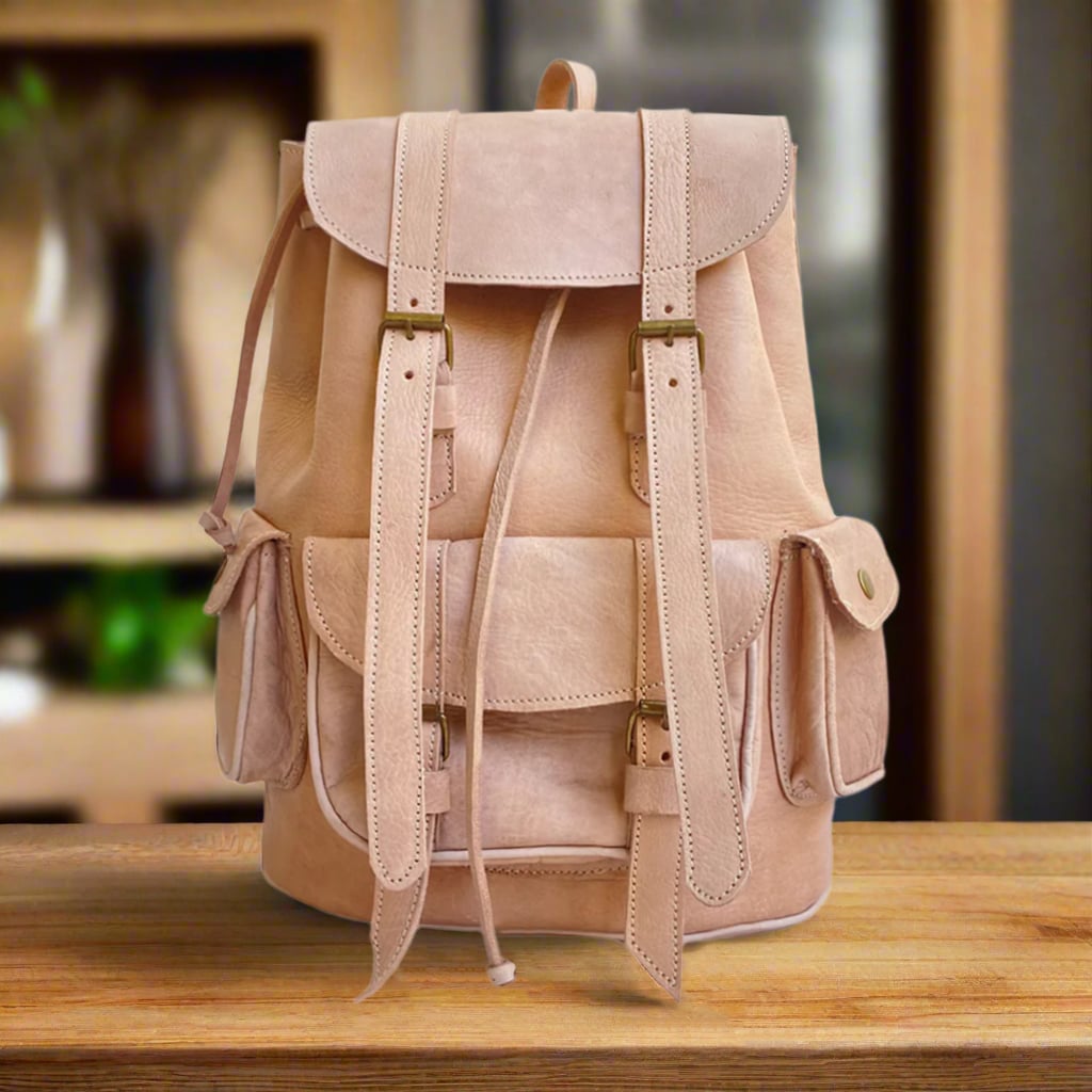 Front view of a handcrafted natural leather backpack with a drawstring and buckle closure, two side pockets, and stitched detailing, displayed on a wooden table with shelves and neutral decor in the background