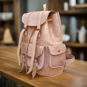 Angled side view of a natural leather backpack featuring buckle straps, spacious side pockets, and a smooth untreated leather finish, set on a wooden table in a warm, rustic interior with pottery and decor