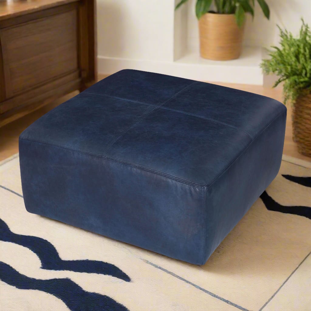 Handcrafted square navy blue leather ottoman in a stylish living room with a beige and navy rug, wooden furniture, and green plants for decor.