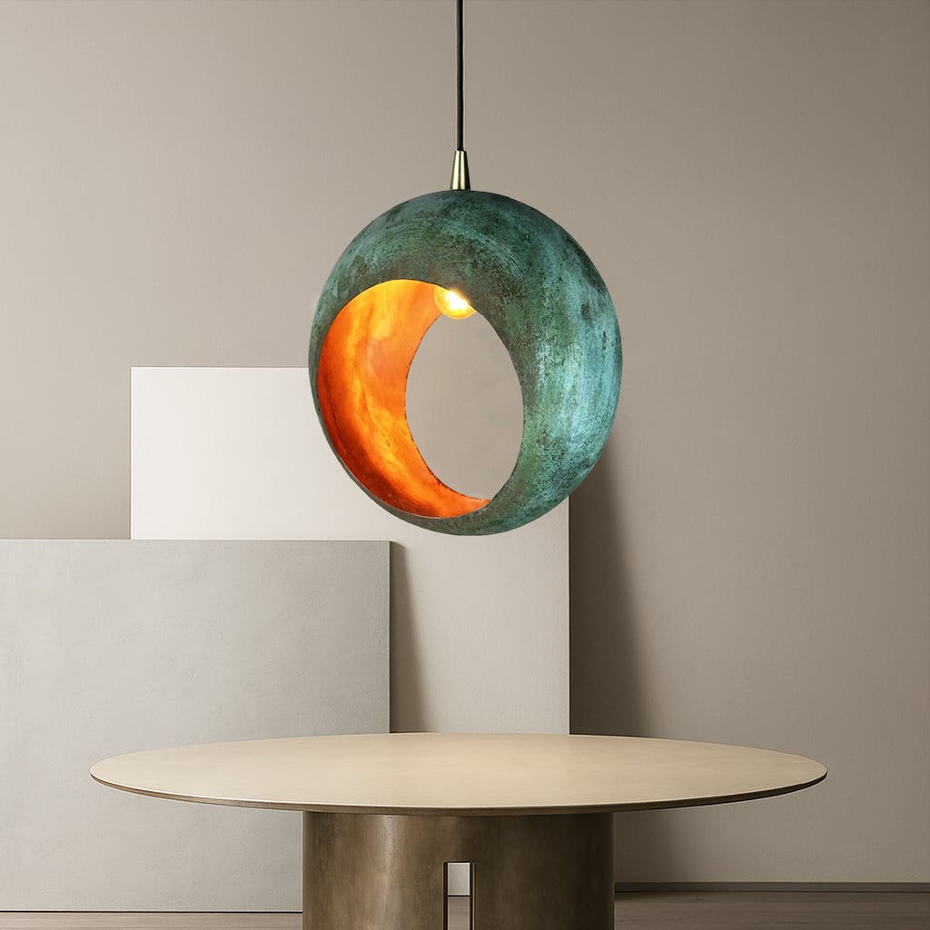 A modern pendant lamp with a spherical, oxidized copper-like exterior and a glowing orange interior, hanging above a minimalist round table in a neutral-toned room