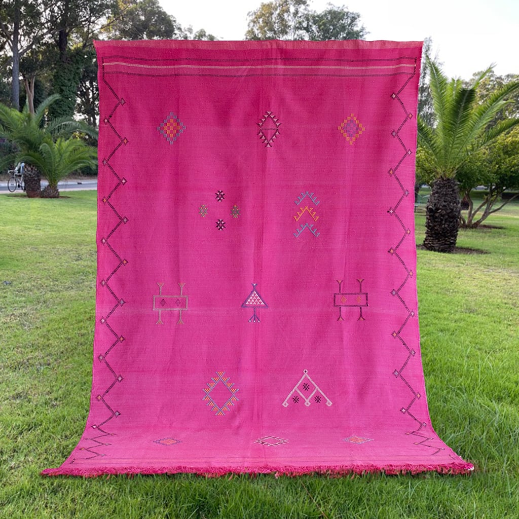 Full view of a bright pink Moroccan rug displayed on grass, adorned with traditional handwoven Berber motifs and bordered with a zigzag pattern