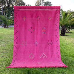 Full view of a bright pink Moroccan rug displayed on grass, adorned with traditional handwoven Berber motifs and bordered with a zigzag pattern