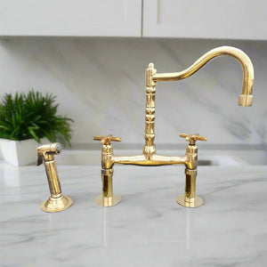 Polished brass bridge kitchen faucet with cross handles and a matching side spray, mounted on a marble counter in a traditional kitchen setting