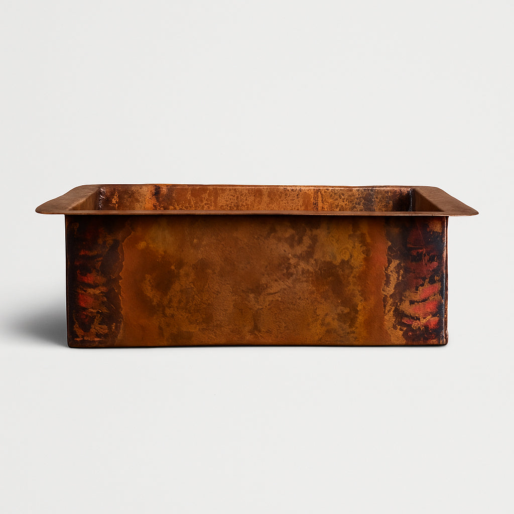 A rectangular copper sink with a rich natural patina, showcasing a blend of earthy browns, oranges, and reddish tones, photographed on a light background