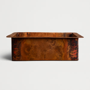 A rectangular copper sink with a rich natural patina, showcasing a blend of earthy browns, oranges, and reddish tones, photographed on a light background
