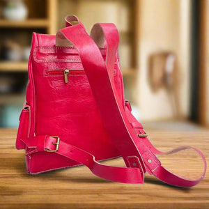 Back view of a red leather and kilim backpack with adjustable leather straps, a secure zippered back pocket, and detailed stitching, set on a wooden surface with soft lighting