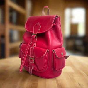 Angled side view of a red Moroccan leather backpack showcasing its structured design, two snap-button side pockets, a large front pocket, and adjustable leather drawstrings under the top flap, set on a wooden table in a softly lit room