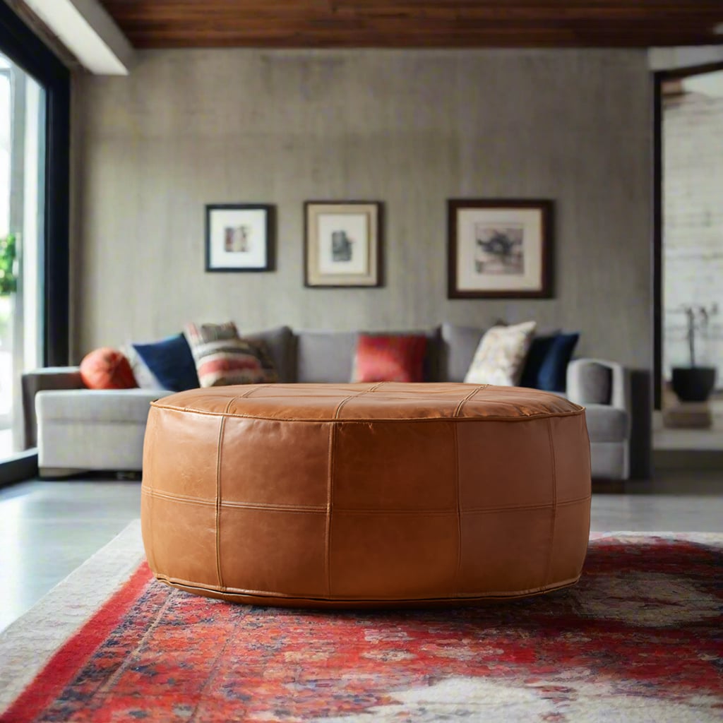 A round tan leather pouf sits on a red patterned rug in a modern living room, with a gray sofa, colorful cushions, framed wall art, and concrete walls in the background