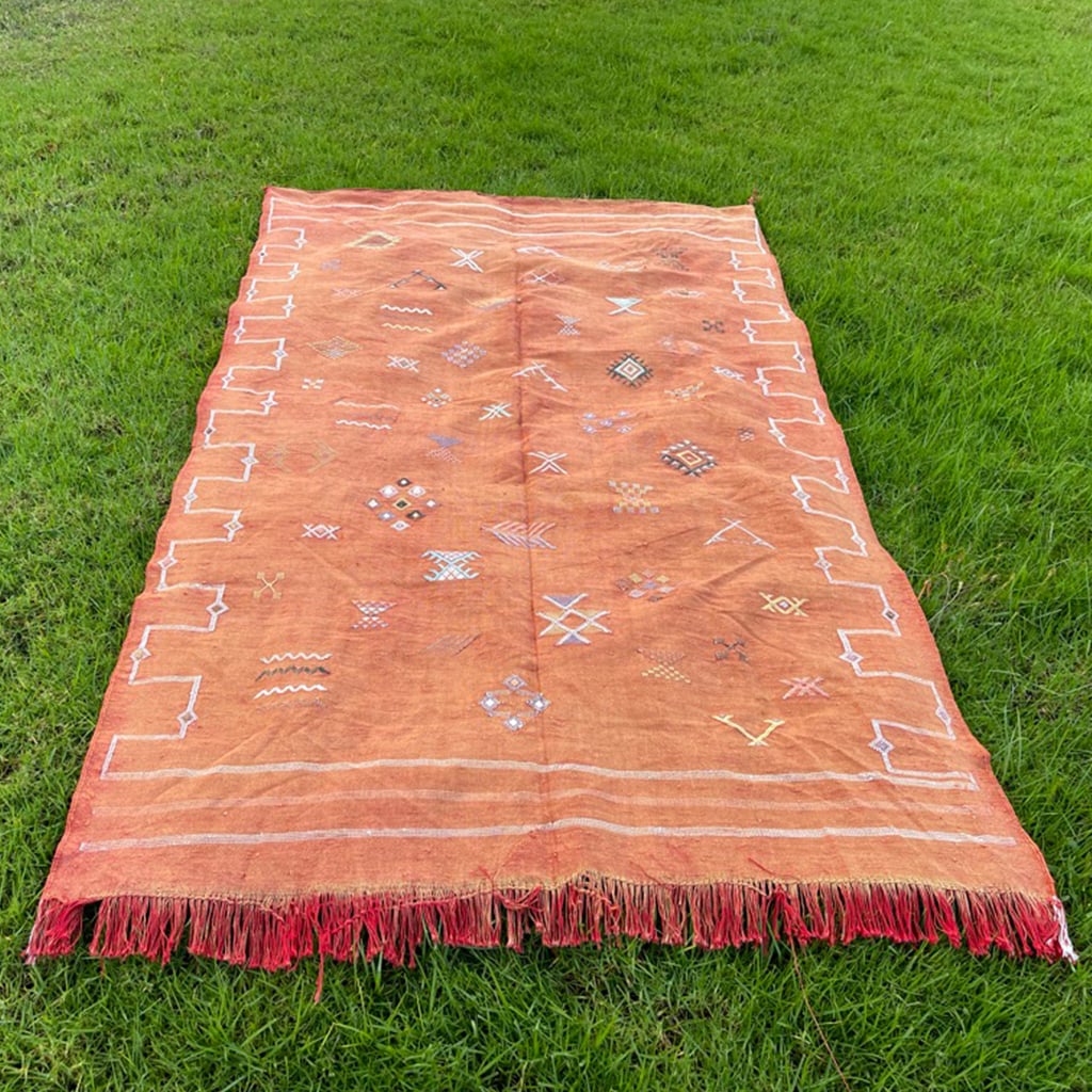 Rust-colored Moroccan rug laid flat on green grass, showing detailed handwoven Berber motifs and bordered by red fringe and white geometric accents