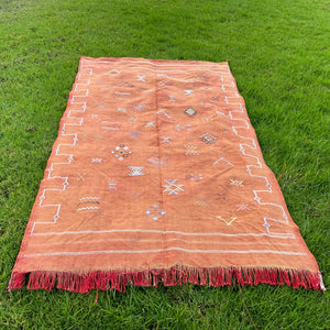 Rust-colored Moroccan rug laid flat on green grass, showing detailed handwoven Berber motifs and bordered by red fringe and white geometric accents