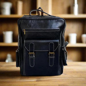 Front view of a sleek black leather backpack featuring a zippered pocket, front buckle pocket, and contrasting white stitching, placed on a wooden surface