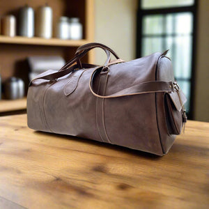 A sleek brown leather duffle bag crafted from premium full-grain leather, featuring a detachable shoulder strap, reinforced handles, and a side pocket. The bag rests on a wooden surface in a softly lit, rustic interior, highlighting its smooth finish and minimalist design