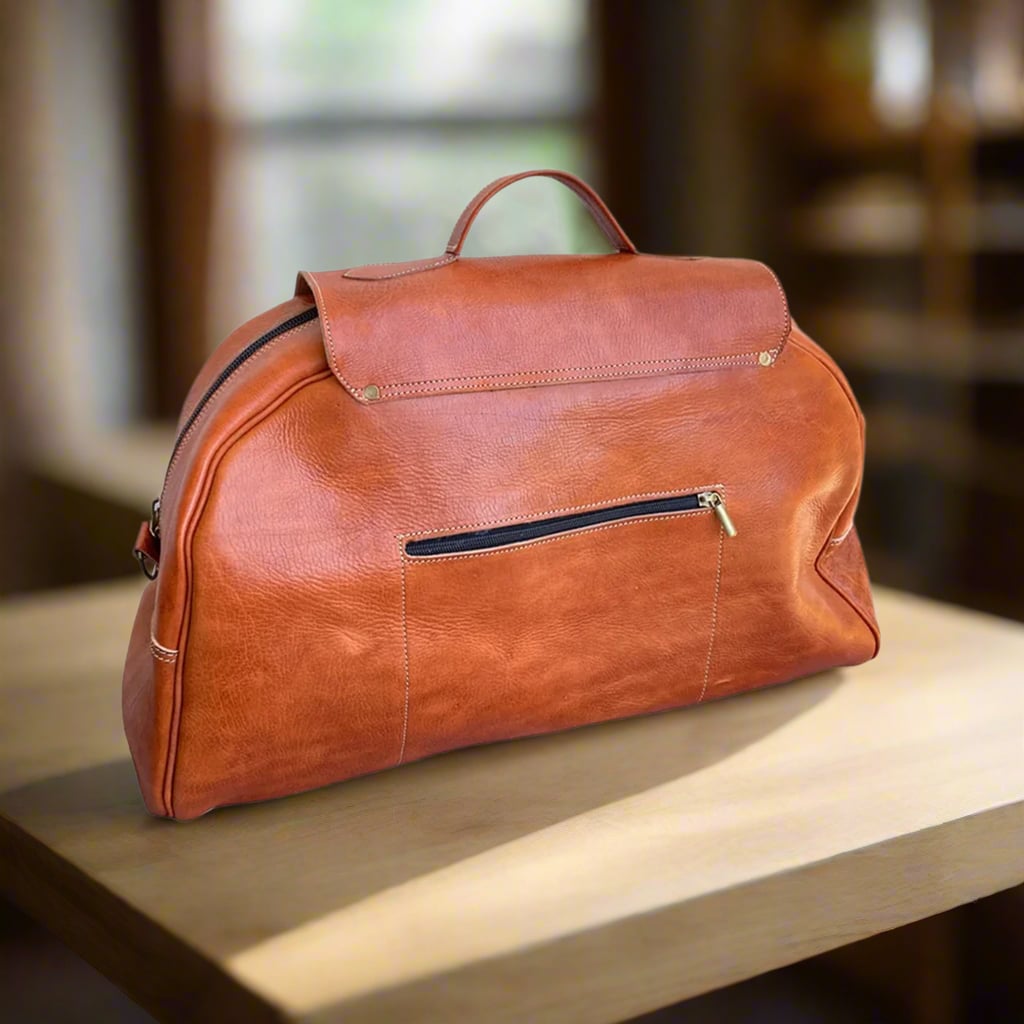 A sleek tan leather travel bag with a minimalist design, featuring a zippered front pocket for easy access and a smooth, durable finish. Set on a wooden table in warm natural light